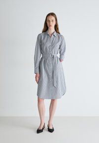 Stripet kjole i blå og hvit, med knapper foran, lange ermer, belted midje og asymmetrisk kant. Teksturert stoff.