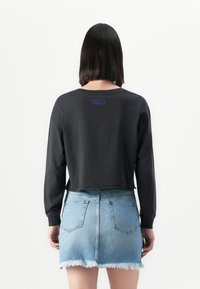 Woman with black long-sleeve top and light blue frayed denim mini skirt, standing facing a white background.