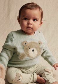 Vaaleanvihreä neulepullover, jossa on teksturoitu karhuaihe; vaaleat housut, joissa on karhuaplikaatiot polvissa. Pehmeät materiaalit, pyöreät muodot.