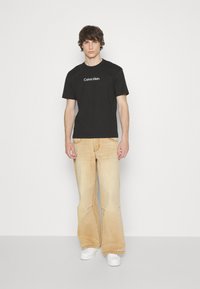 T-shirt en coton noir avec le logo "Calvin Klein" blanc, associé à un jean beige clair évasé et des baskets blanches. Fond uni.