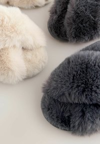 Soft, plush slippers in light beige and dark gray, featuring a closed toe design, thick fur texture, and rubber soles for grip.