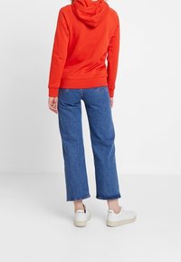 Hoodie en coton rouge avec une capuche à cordon, assorti à un jean en denim bleu à jambes larges et des baskets blanches avec des semelles claires.