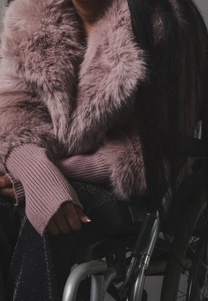 Persoon met lang donker haar in een roze bontjas en vingervrije handschoenen, zittend in een rolstoel met handen op de schoot.