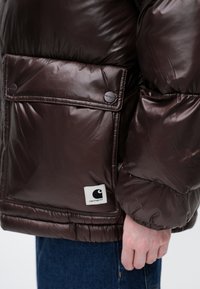 Chaqueta acolchada marrón y brillante con un gran bolsillo frontal, botones a presión y un parche de logo blanco. Textura acolchada y diseño acolchado.