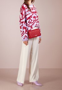 Pestrý svetr s červenými a růžovými vzory, bílé kalhoty s širokými nohavicemi, červená prošívaná kabelka a levandulové lodičky se špičatou špičkou.