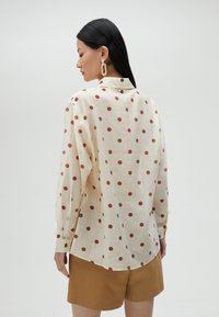 Light-colored shirt with red polka dots, long sleeves, button-up collar, and a rounded hem. Worn with brown shorts, featuring a smooth texture.