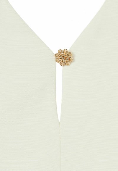 Tejido color crema con un diseño de escote en V profundo, que presenta un cierre metálico dorado en forma de flor en el cuello.
