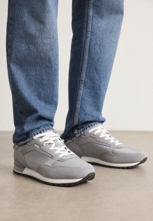 Person iført grå ruskindssneakers med hvide snørebånd og blå jeans, stående på en beige gulv.