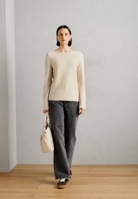 Sweater de malha bege, jeans cinza de perna larga e sapatos castanhos. A segurar uma bolsa branca com uma alça estampada. Textura suave, design moderno.