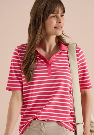 Femme portant un polo à manches courtes rayé rose et blanc, un pantalon beige et portant un sac à épaule beige, regardant sur le côté.