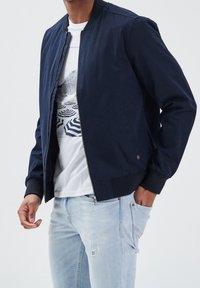 Mörkblå bomberjacka med dragkedja och ribbade ärmslut, bärs över en vit t-shirt med geometrisk design, ihop med ljusblå jeans.