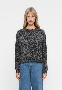 Giovane donna con capelli biondi lisci che indossa una maglietta a maniche lunghe grigio scuro con stampa leopardata e jeans blu larghi, in piedi su uno sfondo bianco.
