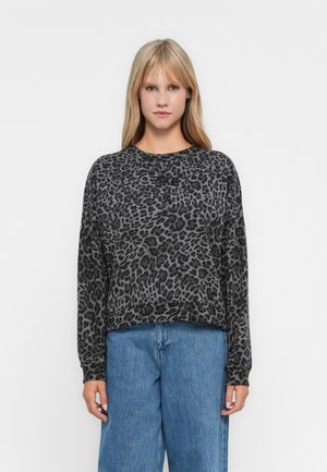 Giovane donna con capelli biondi lisci che indossa una maglietta a maniche lunghe grigio scuro con stampa leopardata e jeans blu larghi, in piedi su uno sfondo bianco.