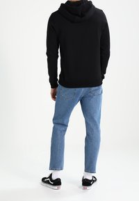 Felpa nera con tasca a marsupio, abbinata a jeans azzurri chiari e sneakers nere con dettagli bianchi, in piedi davanti a uno sfondo neutro.
