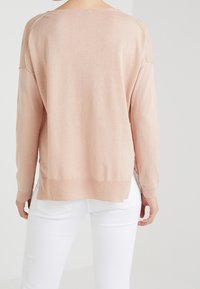 Miękki różowy sweter z dzianiny o długich rękawach, luźnym kroju i rozcięciach po bokach u dołu, w połączeniu z białymi spodniami.