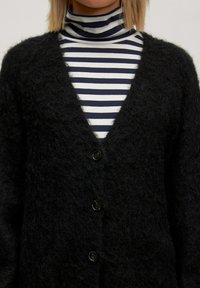 Schwarze, flauschige Strickjacke mit tiefem V-Ausschnitt und fünf Knöpfen, über einem schwarz-weißen gestreiften Rollkragenpullover getragen.