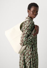 Grande borsa tote off-white realizzata in pelle liscia con un design minimalista e un manico aperto, indossata sulla spalla sopra un vestito a fantasia.
