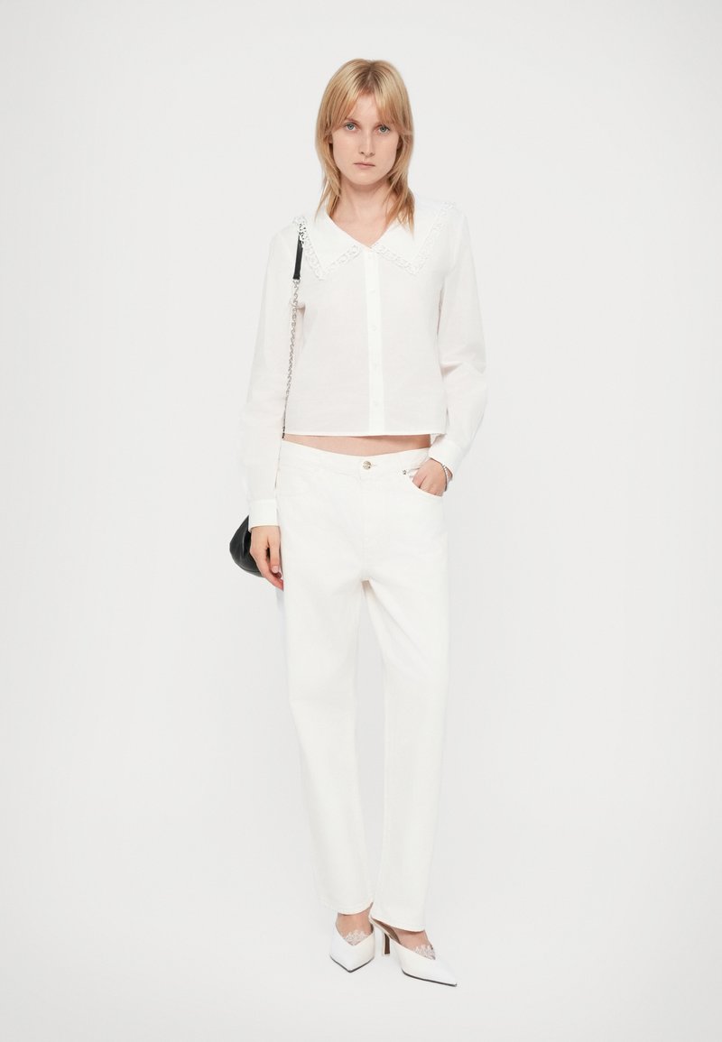 Weißes, verkürztes Hemd mit Knöpfen und Spitzenkragen, kombiniert mit weißen, hoch taillierten Hosen und weißen spitzen Absatzschuhen. Schwarze Schultertasche.