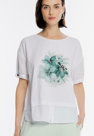 Kobieta ubrana w białą koszulę z krótkim rękawem z turkusowym kwiatowym wzorem i zapinanymi mankietami, w połączeniu z jasnymi spodniami.