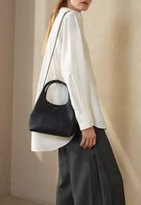 Schwarze Lederhandtasche mit abgerundetem Griff, mit strukturiertem Oberflächen und minimalistischem Design, getragen über einem weißen Hemd und grauen Hosen.