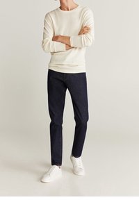 Felpa di colore crema chiaro con texture, jeans slim-fit blu e sneakers bianche. L'outfit è casual e presenta un classico colletto rotondo.