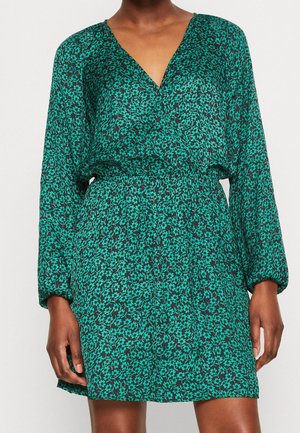 Mujer con un vestido largo de manga larga verde con flores, escote en V y cintura elástica, posando contra un fondo claro y liso.