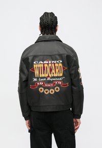 Melna ādas jaka ar daudzkrāsainiem izšuvumiem ar kazino tēmu uz muguras, tostarp uzrakstu "WILDCARD" un dekoratīviem akcentiem.