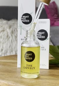Klar glasflaska med lock av trä. Innehåller gul ansiktsolja. Etiketten har en svart cirkel med texten "JAG VILL HA DIG NAKEN" och "DE UNDERBARA."
