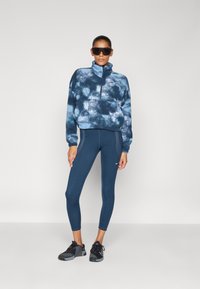 Blå tie-dye fleecepullover med høj krave, lynlåsdetalje, parret med tætsiddende navy leggings og sorte atletiske sko. Store sorte solbriller bæres.