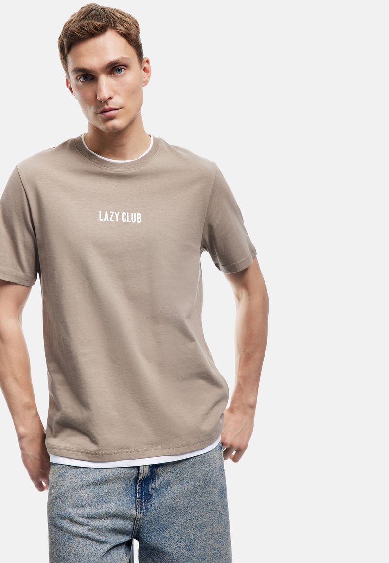 Beige bomulds T-shirt med "LAZY CLUB" trykt i hvid, rund halsudskæring, korte ærmer og en afslappet pasform, parret med lyseblå jeans.