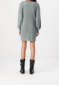 Femme portant une robe-pull en tricot gris clair et des bottes noires mi-mollet avec plusieurs boucles argentées, debout face à un mur blanc.