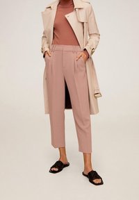 Abrigo trench beige sobre una blusa de canalé color óxido, combinado con pantalones cortos rosa claro y sandalias negras de tipo slide con un patrón texturizado.