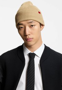 Cappello beige lavorato a maglia con texture a coste e una piccola etichetta rossa con il logo, indossato con una giacca nera testurizzata e una camicia bianca con cravatta nera.