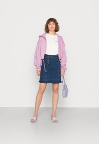 Hoodie roxo, t-shirt branca, saia de ganga escura com cintura atada, mules lavanda e uma pequena mala combinando. Fundo simples.