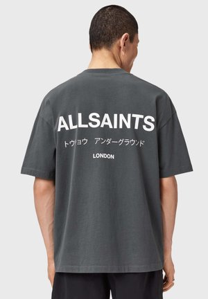 Mężczyzna zwrócony tyłem, ubrany w ciemnoszarą, oversize'ową koszulkę z białym napisem „ALLSAINTS”, japońskimi znakami i napisem „LONDON” na plecach.