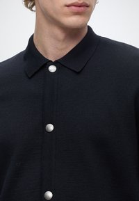Gros plan d'une personne portant une chemise noire à col avec trois boutons-pression argentés visibles sur le devant.