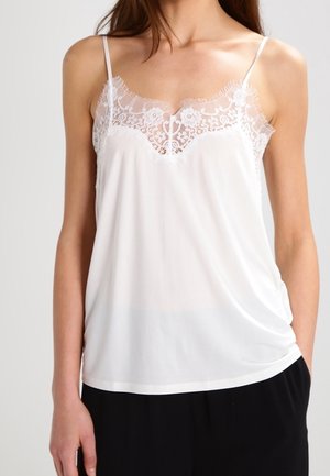 Camiseta blanca sin mangas con detalle de encaje floral en el escote, combinada con pantalones negros, mostrada desde el cuello hasta las caderas, mano izquierda del modelo en el bolsillo.