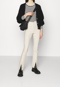 Černý pletený cardigan přes pruhovanou košili s dlouhým rukávem, doplněný krémovými kalhotami s bočními rozparky a černými kotníkovými botami.