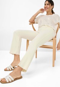 Crème-kleurige flared jeans met raw hems, gecombineerd met lichte slip-on sandalen met een geweven ontwerp, zittend op een houten stoel in mid-century stijl.