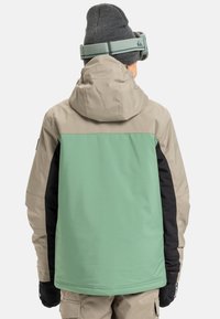 Lichtgewicht, geïsoleerde jack met een mintgroen achterpaneel, een beige bovenstuk en zwarte zijpanelen, voorzien van een capuchon en handschoenen.