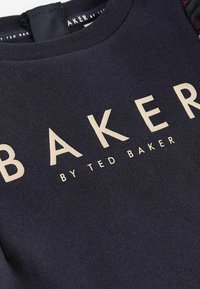 Granatowy sweter z wyraźnym beżowym logo "BAKER" oraz mniejszym napisem "by TED BAKER". Miękki, teksturowany materiał, klasyczny krój z okrągłym dekoltem.