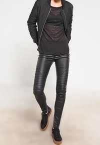 Schwarze Bomberjacke über einem transparenten schwarzen Top, kombiniert mit enganliegenden schwarzen Kunstlederhosen mit Rippendetails und schwarzen Sneakers.