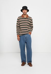 Langsleeve polo shirt met groene, witte en bruine strepen, bruine kraag; gecombineerd met losse blauwe jeans en bruine leren schoenen. Zwarte bucket hat.