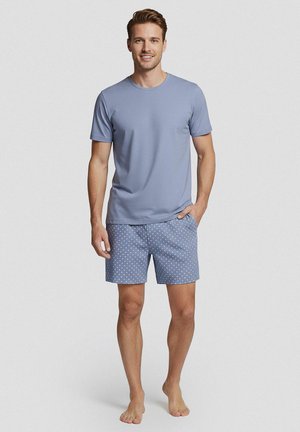 Man staat op blote voeten, draagt een lichtblauw T-shirt met korte mouwen en blauwe korte broek met wit gestippeld patroon op een effen achtergrond.