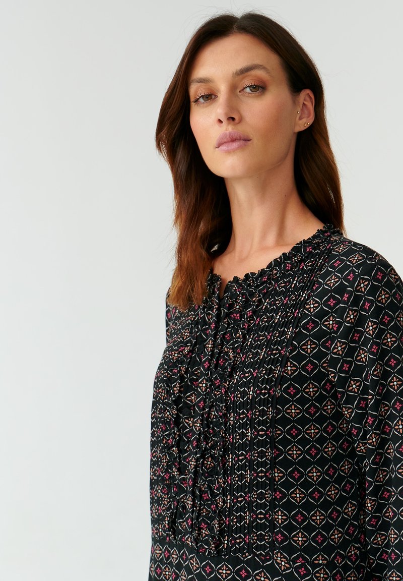 Czarna bluzka z kwiatowym wzorem geometrycznym w odcieniach czerwieni, różu i kremu. Posiada plisowane detale oraz falbaniaste wykończenie dekoltu, wykonana z miękkiego materiału.