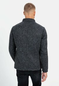 Grijze jas met textuur en een blauw gespikkeld patroon, met een staande kraag en een getailleerde pasvorm, gezien van de achterkant met donkere spijkerbroek.