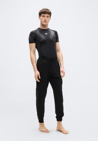 Μαύρο αθλητικό μπλουζάκι με κοντά μανίκια, διακοσμημένο με υφασμάτινες λεπτομέρειες και λογότυπο. Συνδυάζεται με μαύρο παντελόνι φόρμας με ελαστικά περιλαίμια και πλαϊνές τσέπες.