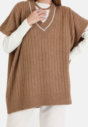 Kvinde iført oversized brun ribstrikket ærmeløs sweater over hvid langærmet top, kombineret med hvide bukser og en chunky guldkæde.