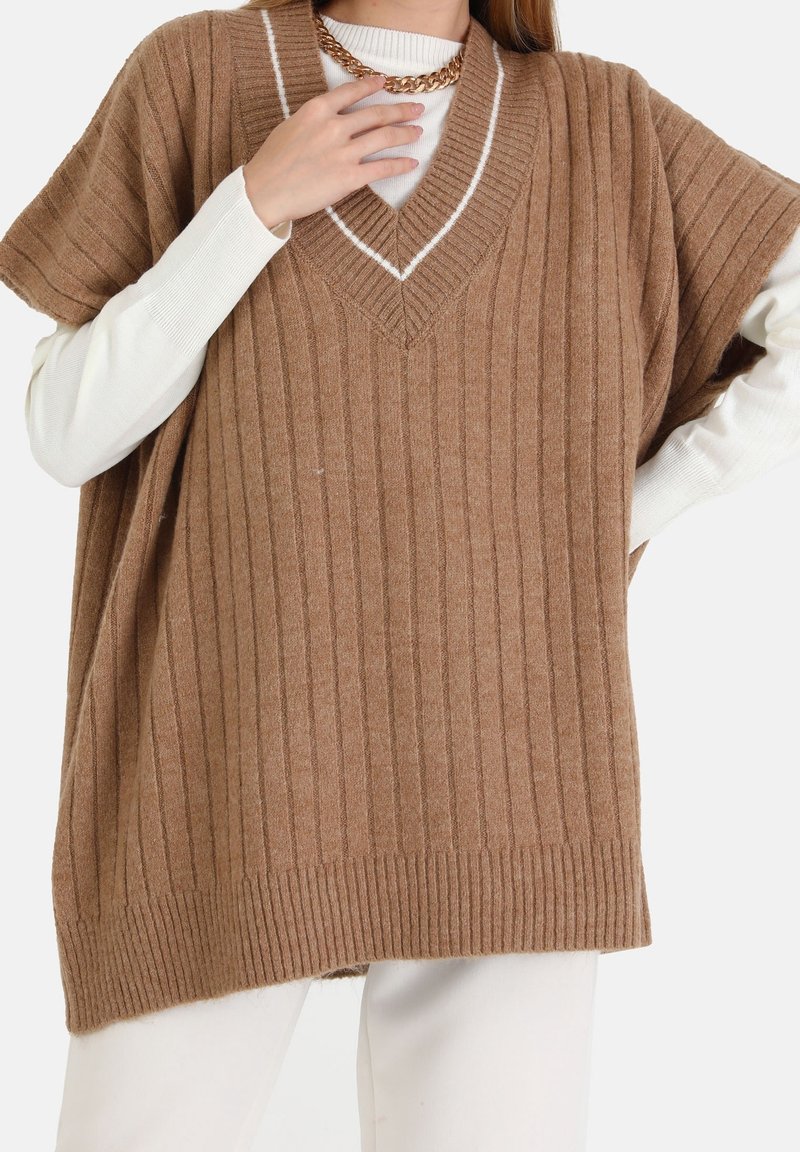 Femme portant un pull sans manches marron côtelé oversize sur un haut blanc à manches longues, associé à un pantalon blanc et un collier chaîne doré épais.