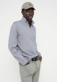 Ανοιχτό γκρι φούτερ με φερμουάρ, μακριά μανίκια και ριμπ τελείωμα, σετ με ανοιχτό πράσινο παντελόνι και μπεζ καπέλο. Διακριτικό λογότυπο.